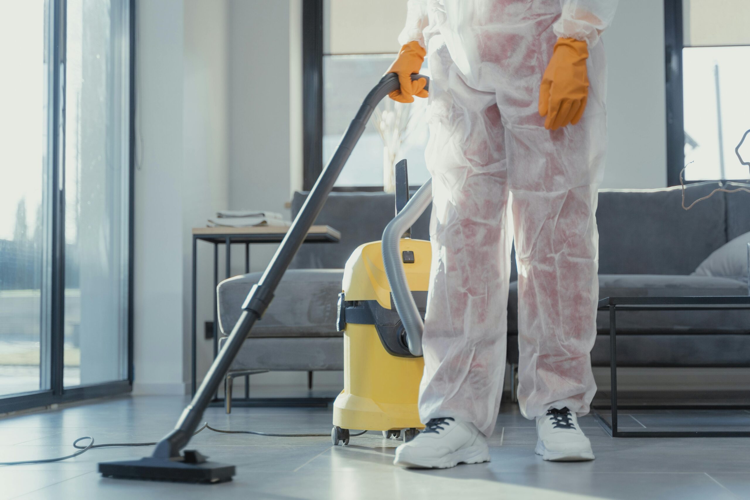 Services A cleaner in protective coveralls and gloves vacuuming a modern living room.